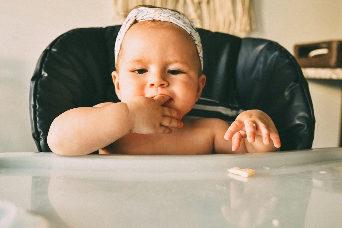 Baby sitzt im Hochstuhl am Tisch und isst selbststaendig mit den Haenden